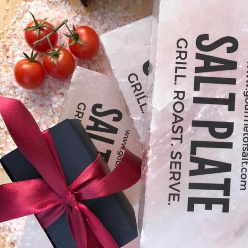 Op de afbeelding: Een cadeaudoos ingepakt in zwart papier met een rood satijnen lint ligt op een roze Himalaya-zoutplaat. De zoutplaat is gelabeld met "SALT PLATE" en "GRILL. ROAST. SERVE." met het websiteadres "www.gourmetsalt.com".