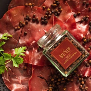 Op de afbeelding: Een glazen pot gevuld met rode peperkorrels ligt op een bed van dun gesneden gedroogd vlees. De pot heeft een rood etiket met de tekst "Salt's Up" en "Kampot Red Pepper" erop gedrukt.