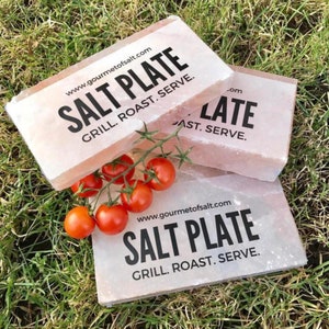 Puede incluir: Tres placas de sal rosa del Himalaya con el texto "www.gourmetofsalt.com SALT PLATE GRILL. ROAST. SERVE." impreso en ellas. Las placas están sobre un lecho de hierba verde.