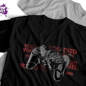 Puede incluir: Camiseta negra con un gráfico de un mando de videojuego roto y el texto "You Died" repetido varias veces en rojo.
