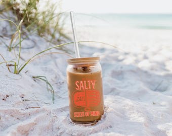 May include: A glass jar with a bamboo lid and a clear straw, filled with a brown liquid. The jar has a graphic that says "The Salty State of Mind" with a stylized image of a seashell.