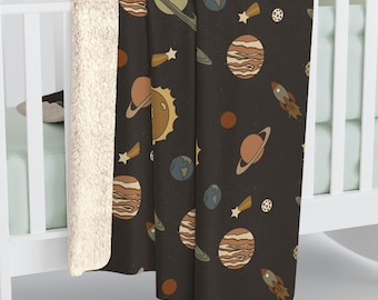 May include: A black blanket with a repeating pattern of planets, stars, and rockets in shades of brown, tan, and blue. The blanket is draped over a white crib, with a cream-colored, fluffy backing visible.