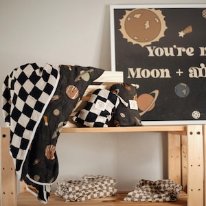 Puede incluir: Una manta de bebé a cuadros en blanco y negro con un estampado de temática espacial. La manta está doblada y se muestra en un estante de madera con un letrero que dice "You're my Moon + all my Stars".