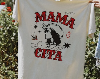 May include: Cream-colored t-shirt featuring the words "MAMA CITA" in red, stylized lettering. The design includes a woman in a cowboy hat, stars, and other decorative elements. The shirt is held up in front of a green, leafy background.