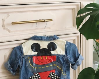 May include: A blue denim jacket with a white and red Mickey Mouse design on the back. The design features Mickey Mouse driving a race car with checkered flags.