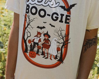 May include: Cream-colored t-shirt with a Halloween-themed graphic. The design features the words "Let's BOO-GIE" in a retro font, surrounded by a border with bats, trees, and children in costumes. The children are holding jack-o'-lanterns.