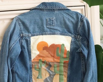 May include: A blue denim jacket with a painted desert scene on the back. The scene features a sunset, mountains, and two cacti.