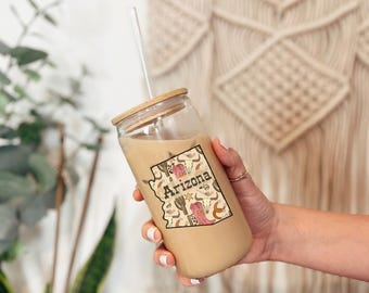 May include: A clear glass tumbler with a wooden lid and straw, filled with a beige beverage. The tumbler features a sticker with the outline of Arizona, filled with desert-themed illustrations and the state name.