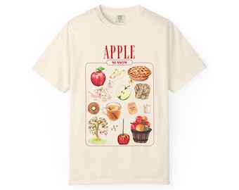 May include: Cream-colored t-shirt featuring a watercolor illustration of apple-themed items. The design includes a red apple, apple slices, a pie, a donut, a cup of tea, and a basket of apples. The text "APPLE SEASON" is at the top.