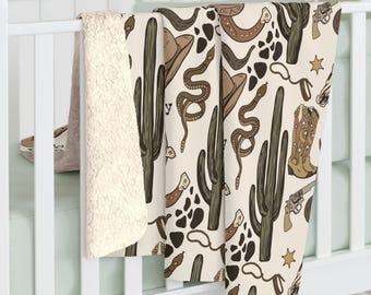 May include: A cream-colored baby blanket with a repeating pattern of cowboy-themed illustrations. The design includes cacti, cowboy hats, snakes, boots, and revolvers in shades of brown and tan. The blanket is displayed over a crib.