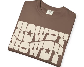 May include: A brown t-shirt with the word "HOWDY" in a cream-colored, retro font. The word is stacked twice, with stars replacing the "O" in each word. The shirt is folded and has a tag in the upper right corner.