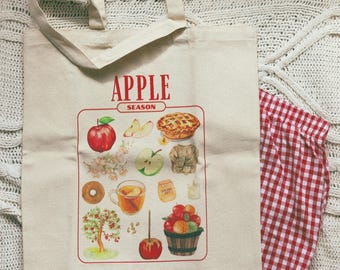 May include: A beige tote bag with the words "APPLE SEASON" in red, featuring illustrations of apples, apple pie, and other apple-related items. A red and white checkered fabric is visible.