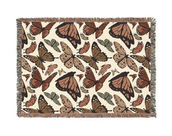 May include: A rectangular woven throw blanket featuring a repeating pattern of butterflies in shades of brown, orange, and green on an off-white background. The blanket has a fringed edge.