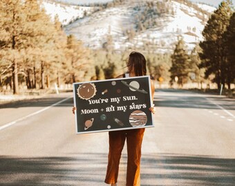 May include: A rectangular sign with a black background and white border, featuring the text "You're my Sun, Moon + all my stars." The sign is decorated with illustrations of planets and celestial bodies. The person holding the sign is wearing brown pants and white sneakers.