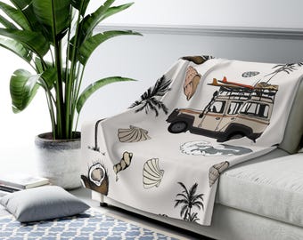 May include: A white throw blanket featuring a repeating pattern of a vintage off-road vehicle with a surfboard, palm trees, seashells, and coconuts. The blanket is draped over a sofa, with a large potted plant in the background.