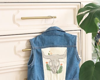 May include: A blue denim vest with a white patch on the back featuring a line drawing of a cow skull with cacti and flowers.