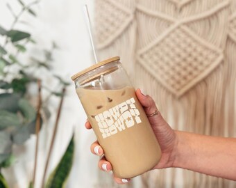May include: A clear glass tumbler filled with iced coffee, featuring a wooden lid and a white straw. The tumbler has the words "HOWDY HOWDY" in a wavy, retro font. The glass is held by a person with a gold ring.
