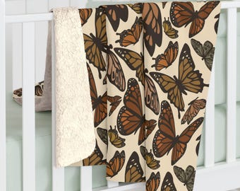 May include: A cream-colored blanket with a repeating pattern of brown and orange butterflies. The blanket has a soft, sherpa-like backing. The butterflies vary in size and are in different stages of flight.