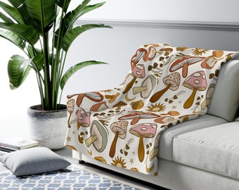 May include: A cream-colored throw blanket featuring a repeating pattern of various mushrooms in shades of brown, pink, and green. The blanket is draped over a white couch, creating a cozy and inviting aesthetic. The design includes suns and stars.