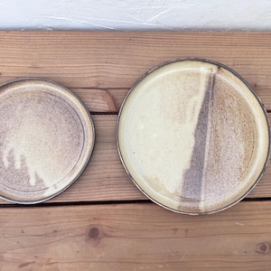 May include: Two ceramic plates with a speckled, earthy design. The plates have a neutral colour palette with shades of beige, brown, and cream. The plates are round with a slightly raised edge. The plates are on a wooden surface.