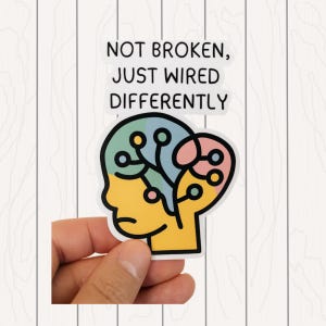 May include: A sticker with the text "NOT BROKEN, JUST WIRED DIFFERENTLY" above a colorful illustration of a human head with a brain-like design. The head is yellow, the brain is blue and pink.