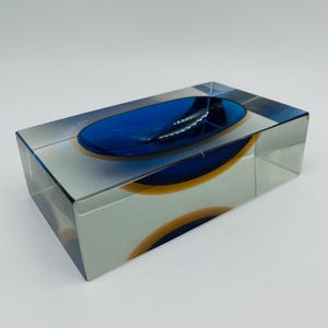 May include: A rectangular glass ashtray with a deep blue oval recess. The ashtray features an orange and blue design within the clear glass, creating a layered effect. The design is modern and geometric in style.
