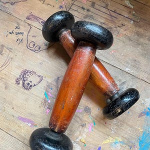 May include: Two vintage wooden dumbbells with black painted ends and a reddish-brown handle. The dumbbells are resting on a wooden surface with paint splatters and markings. The dumbbells appear to be antique and well-used.