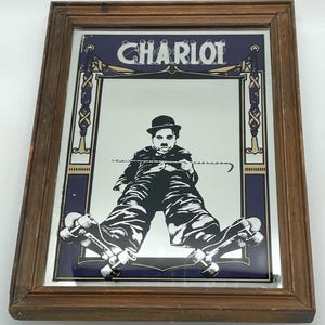 May include: A framed mirror with a black and white image of Charlie Chaplin on roller skates. The word "CHARLOT" is printed in white on a dark blue background above the mirror. The frame is made of brown wood.