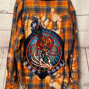 May include: Orange and blue plaid flannel shirt with a tie-dye patch featuring a skeleton hand holding a rose and the text "GD".