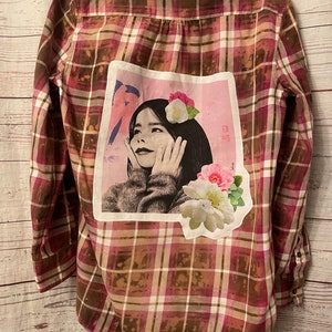 May include: A brown and pink plaid flannel shirt with a white patch sewn on the back. The patch features a black and white image of a woman with flowers and Japanese text.