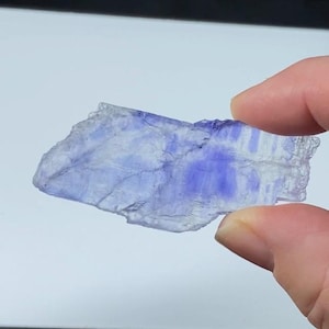 Puede incluir: Un espécimen mineral translúcido de forma irregular con una mezcla de tonos azul claro y púrpura intenso. La piedra se sostiene entre los dedos, mostrando su textura natural y cruda y sus variaciones de color. El mineral parece ser un tipo de fluorita.