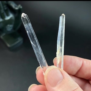 May include: Two clear, elongated crystal formations are held in a hand. The crystals are transparent with pointed tips and a slightly rough texture. The background is dark, emphasizing the crystals' clarity.