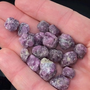 May include: A handful of raw ruby gemstones, displaying a deep pink-red hue. The stones have a rough, natural texture. The image shows the gemstones held in a hand, highlighting their unpolished state. Suitable for jewellery making or as collectibles.