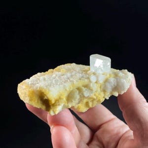 Puede incluir: Una formación cristalina de color claro con una estructura cristalina prominente y vertical. La base es una mezcla de amarillo y blanco, con una superficie rugosa y texturizada. El fondo es negro.