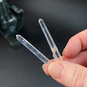 May include: Two clear quartz crystal points with a pale pink hue at the tips. The crystals are held in a hand against a dark background. The crystals are transparent and have a smooth, polished appearance.