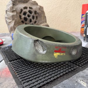 May include: An olive green, circular object with a red and yellow design, possibly a component of a larger structure. The object sits on a black, textured mat. Paint cans and a stone-like structure are in the background.