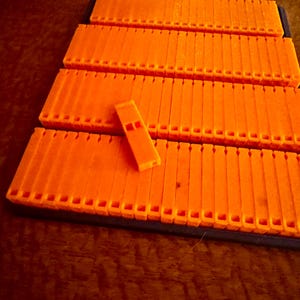 May include: Bright orange plastic components arranged in a stepped configuration. The components have a series of vertical slots and a small rectangular piece with two holes. The items are on a dark surface.