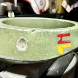 May include: A green, round, ceramic bowl with a red and yellow "H" painted on the side. The bowl has a chipped edge and a small hole on the side.