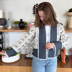 May include: A quilted jacket with a dragonfly pattern on a cream background, accented with navy blue panels and white trim. The jacket is paired with light blue jeans. The jacket's design is a blend of patterns and colors.