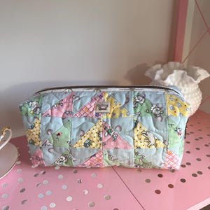 May include: A rectangular quilted cosmetic bag with a zipper closure. The bag features a patchwork design with pastel triangles and a repeating pattern of cartoon animals. The text "Quilting Quail" is visible on the bag.