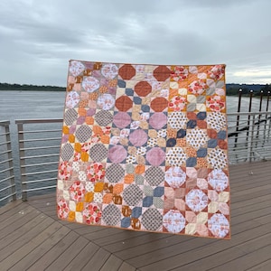 May include: A handmade quilt with a patchwork design. The quilt features a variety of fabrics in shades of orange, pink, brown, and white. The pattern includes circles, squares, and floral prints. The quilt is displayed outdoors on a wooden deck.