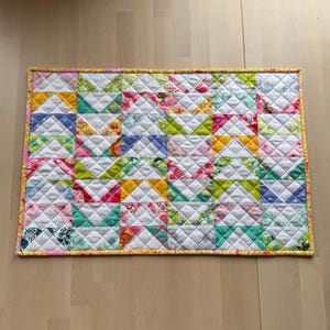 Puede incluir: Un edredón rectangular de patchwork con un borde amarillo. El edredón está hecho de varias telas estampadas en rosa, verde, azul y amarillo, dispuestas en un patrón geométrico. El edredón parece estar hecho a mano.
