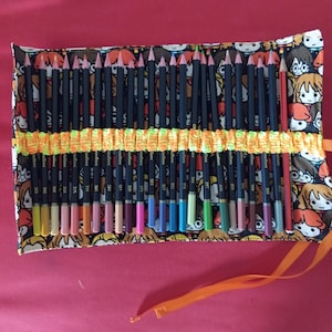 Op de afbeelding: Een stoffen potloodrol met een Harry Potter-thema. De rol is zwart met een kleurrijk patroon van Harry Potter-personages. De rol is open en bevat 24 kleurpotloden. De potloden zijn in een rij gerangschikt en zijn genummerd.