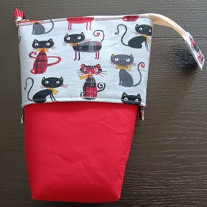 Op de afbeelding: Een rode en grijze stoffen etui met een kattenpatroon. Het bovenste gedeelte heeft een lichtgrijze stof met zwarte katten met vlinderdassen en geruite accenten. De onderkant is effen rood en de etui heeft een ritssluiting en een stoffen lus.