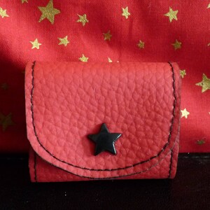 Puede incluir: Una pequeña cartera de cuero roja con un diseño de estrella negra. La cartera tiene un cierre de solapa y un borde negro cosido.