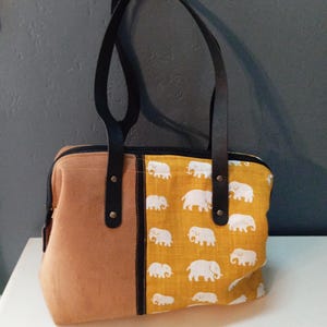 Peut inclure: Sac fourre-tout beige et jaune avec des poignées noires. Le sac présente un matériau beige semblable à du daim d'un côté et un tissu jaune avec des motifs d'éléphants blancs de l'autre. Le sac a des poignées en cuir noir et une fermeture éclair noire.
