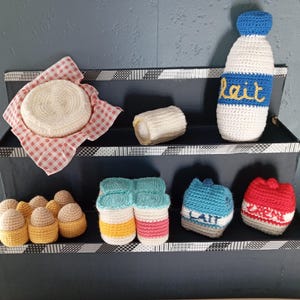 Könnte beinhalten: Eine gehäkelte Präsentation von Lebensmitteln, darunter ein runder Käse auf einem rot-weiß karierten Tuch, eine Milchflasche mit dem Text "Lait" und verschiedene andere gehäkelte Milchprodukte. Die Präsentation befindet sich auf einem schwarzen, mehrstufigen Regal.