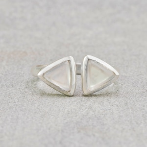 Puede incluir: Anillo de plata con dos triángulos abiertos, cada uno con una incrustación nacarada. Los triángulos están delineados en plata, creando un diseño moderno y geométrico. El anillo se presenta sobre un fondo gris neutro.