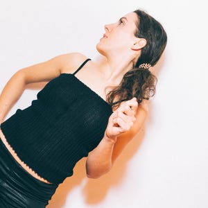 May include: A person wearing a black ribbed tank top with spaghetti straps and black pants. The person is leaning against a white wall, looking upwards. The hair is pulled back in a ponytail.