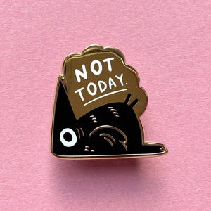 May include: A black enamel pin depicting a stylized cat with a gold-coloured cloud-shaped speech bubble. The text "NOT TODAY" is written in white on the cloud. The pin has a gold-coloured border and is set against a pink background.